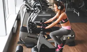 Uśmiechnięta kobieta w słuchawkach odbywa trening na rowerze stacjonarnym dla początkujących.