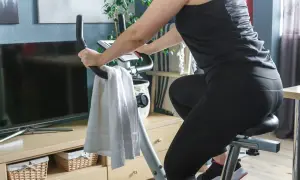 Kobieta ćwiczy na rowerze stacjonarnym. Widać jej nogi w czarnych legginsach i ręce na kierownicy. Obok wisi biały ręcznik. Jazda na rowerze stacjonarnym efekty po miesiącu są widoczne!