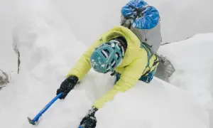 Alpinista w żółtej kurtce wspina się po śnieżnej grani, używając czekana do stabilizacji.