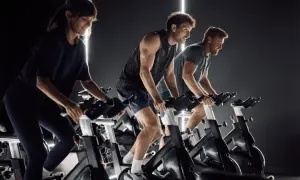 Trzy osoby intensywnie pedałują na rowerach stacjonarnych, w rytmie muzyki, podczas zajęć spinning.