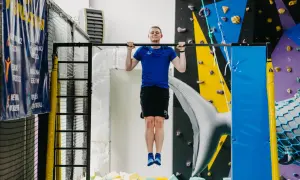 Młody mężczyzna wykonuje ćwiczenia na drążku. W tle ściana wspinaczkowa i basen z gąbkami.