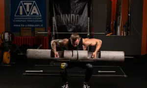 Siłacz przygotowuje się do podniesienia ciężkiej sztangi typu log lift, co jest elementem jego periodyzacji treningowej.