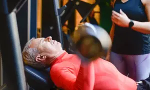 Starszy mężczyzna wykonuje trening funkcjonalny ćwiczenia z hantlem na ławce. Obok stoi kobieta, która go asekuruje.