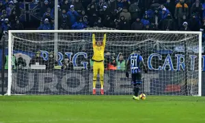 Bramkarz w żółtym stroju wisi na poprzeczce bramki, gotowy do obrony. Naprzeciwko niego, na polu karnym, stoi napastnik z piłką.