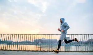 Mężczyzna w bluzie z kapturem uprawia jogging nad rzeką o zachodzie słońca. To świetny sposób na aktywność fizyczną.