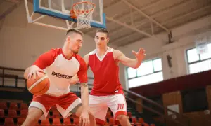 Dwóch koszykarzy w czerwonych strojach na boisku. Jeden z piłką, drugi w obronie. Gra toczy się na podwójnym boisku.