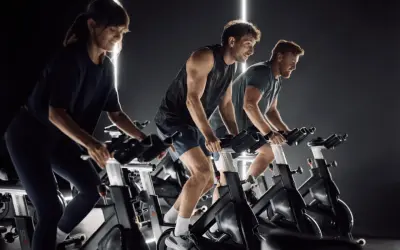 Trzy osoby intensywnie pedałują na rowerach stacjonarnych, w rytmie muzyki, podczas zajęć spinning.