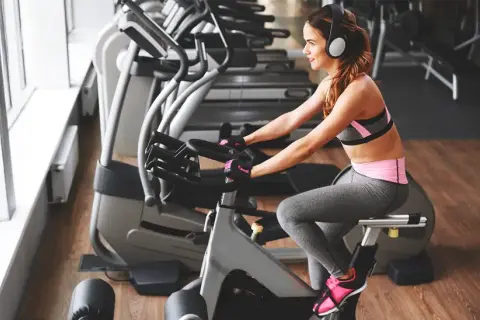 Uśmiechnięta kobieta w słuchawkach odbywa trening na rowerze stacjonarnym dla początkujących.