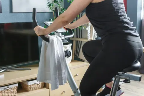 Kobieta ćwiczy na rowerze stacjonarnym. Widać jej nogi w czarnych legginsach i ręce na kierownicy. Obok wisi biały ręcznik. Jazda na rowerze stacjonarnym efekty po miesiącu są widoczne!
