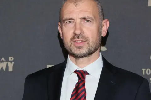 Mężczyzna w garniturze i krawacie, z siwiejącymi włosami. Wygląda jak trener siatkówki mężczyzn, gotowy do analizy gry.