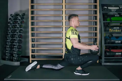 Mężczyzna wykonuje ćwiczenia mobilizacyjne z kettlebell, pogłębiając wykrok. W tle stojaki z hantlami i fitbagami.