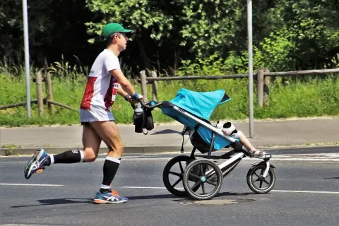 Mężczyzna w zielonej czapce i sportowym stroju bierze udział w biegu z wózkiem. Dziecko w wózku ma na sobie białe skarpetki i sandałki.