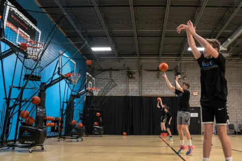 Młody koszykarz trenuje rzuty w hali sportowej, ćwicząc technikę przy użyciu maszyny do podawania piłek.