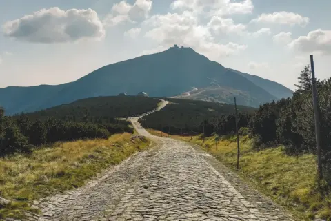 Kamienista droga wije się ku górze Śnieżka, gdzie widać budynki. Wokół zieleń i chmury na niebie.