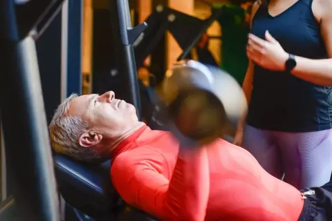 Starszy mężczyzna wykonuje trening funkcjonalny ćwiczenia z hantlem na ławce. Obok stoi kobieta, która go asekuruje.