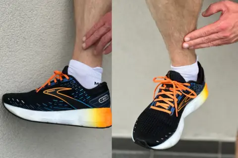 Biegacz pokazuje ból w łydce, który może być objawem shin splints. Na nogach ma czarne buty sportowe Brooks z pomarańczowymi sznurówkami.
