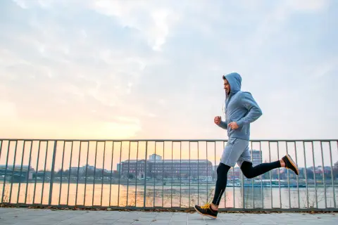 Mężczyzna w bluzie z kapturem uprawia jogging nad rzeką o zachodzie słońca. To świetny sposób na aktywność fizyczną.