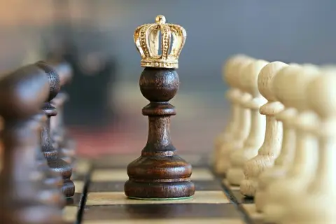Szachownica z figurami. Centralnie ustawiony ciemny pionek z koroną, symbolizujący gambit królowej.