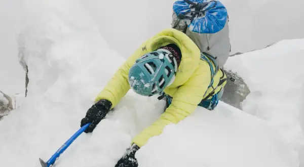 Alpinista w żółtej kurtce wspina się po śnieżnej grani, używając czekana do stabilizacji.