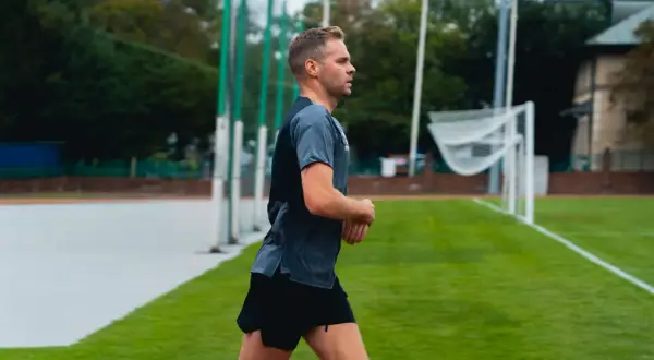 Mężczyzna w sportowym stroju biegnie po bieżni. W tle widać boisko z bramką i drzewa. bc1 bieganie.
