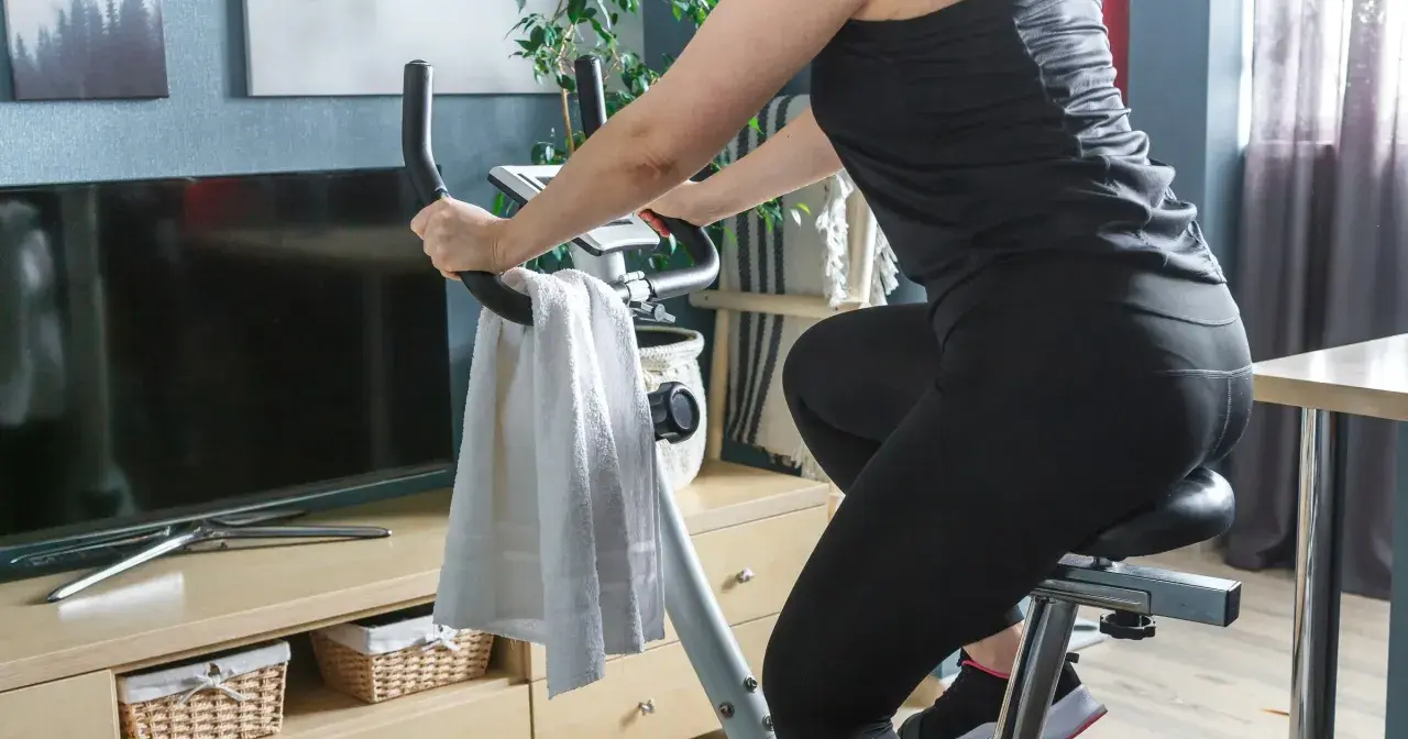 Kobieta ćwiczy na rowerze stacjonarnym. Widać jej nogi w czarnych legginsach i ręce na kierownicy. Obok wisi biały ręcznik. Jazda na rowerze stacjonarnym efekty po miesiącu są widoczne!