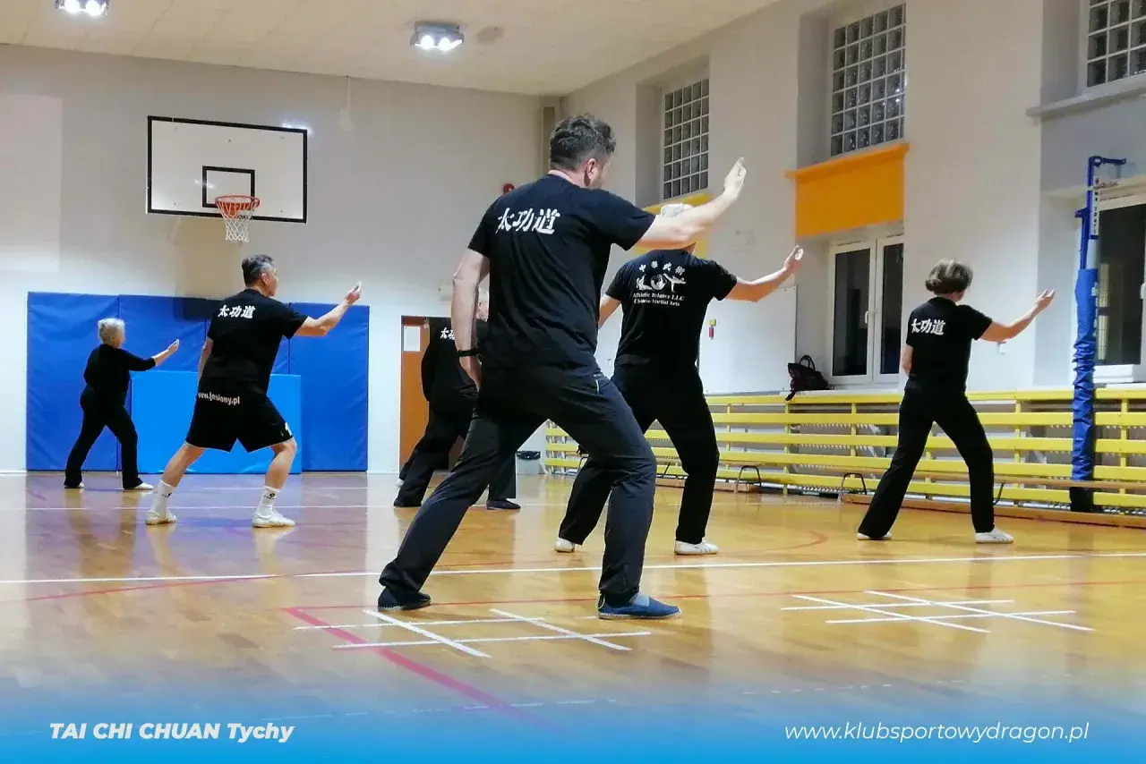 Grupa ćwiczy tai chi w sali gimnastycznej. Wszyscy mają czarne stroje. Warto pamiętać o tai chi przeciwwskazania, choć ćwiczenia są łagodne.