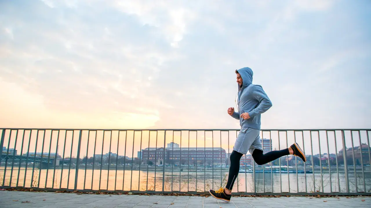 Mężczyzna w bluzie z kapturem uprawia jogging nad rzeką o zachodzie słońca. To świetny sposób na aktywność fizyczną.