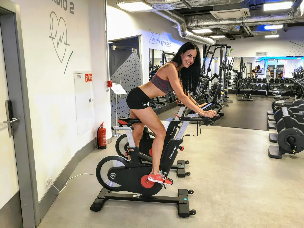 Uśmiechnięta kobieta ćwiczy na rowerze stacjonarnym w siłowni. To idealne miejsce na trening cardio, czy kardio, jak kto woli.