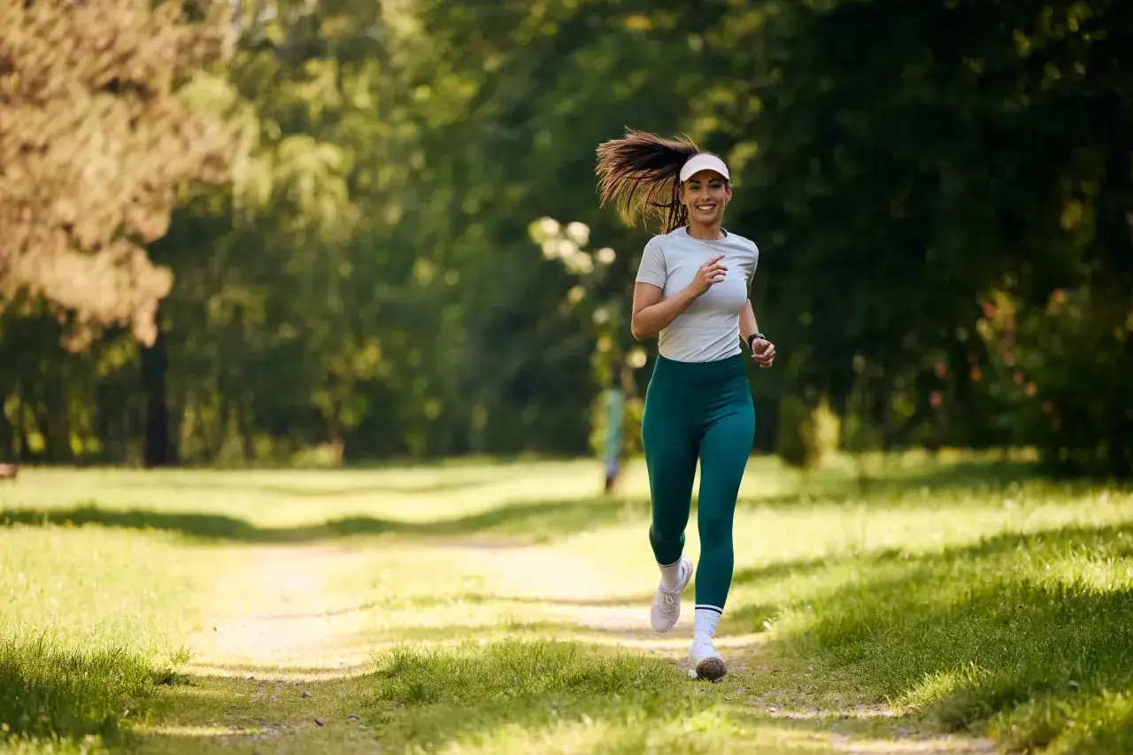 Uśmiechnięta kobieta w sportowym stroju biegnie przez park. To jej bieg na 5 km, czas amatora, ale radość z aktywności jest bezcenna.