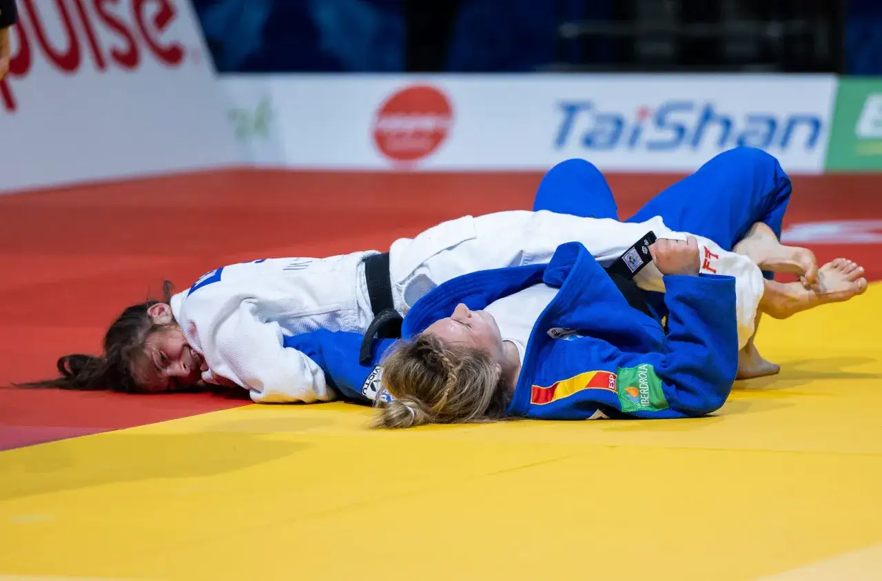Dwie zawodniczki judo leżą na macie po akcji. Jedna z nich, w białym judogi, ma wykrzywioną twarz, sugerując ból lub porażkę. Druga, w niebieskim judogi, leży spokojniej. To mógł być nokaut w judo.