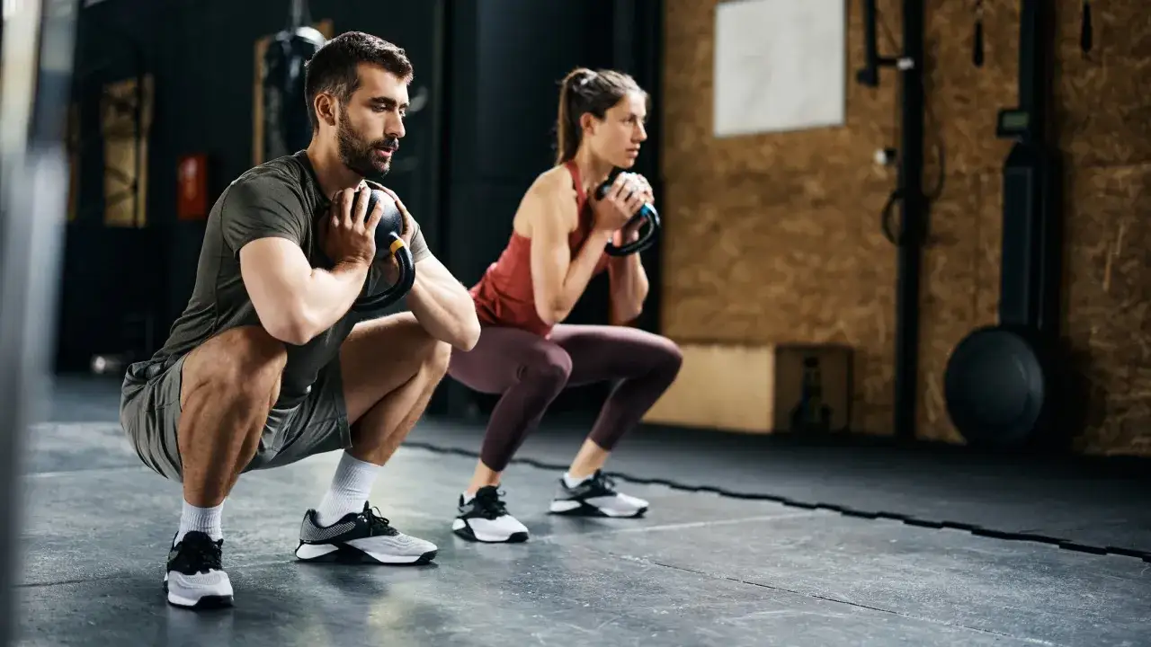 Para ćwiczeń na ból kręgosłupa: mężczyzna i kobieta wykonują przysiady z kettlebellami, wzmacniając mięśnie tułowia.