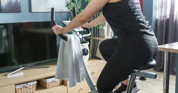 Kobieta ćwiczy na rowerze stacjonarnym. Widać jej nogi w czarnych legginsach i ręce na kierownicy. Obok wisi biały ręcznik. Jazda na rowerze stacjonarnym efekty po miesiącu są widoczne!