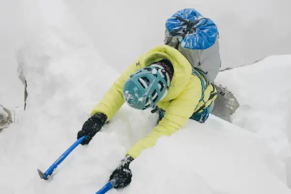 Alpinista w żółtej kurtce wspina się po śnieżnej grani, używając czekana do stabilizacji.