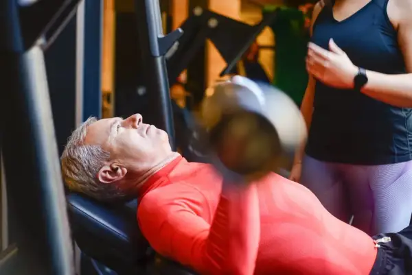 Starszy mężczyzna wykonuje trening funkcjonalny ćwiczenia z hantlem na ławce. Obok stoi kobieta, która go asekuruje.