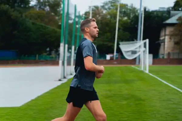 Mężczyzna w sportowym stroju biegnie po bieżni. W tle widać boisko z bramką i drzewa. bc1 bieganie.