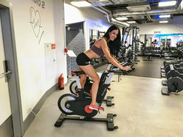 Uśmiechnięta kobieta ćwiczy na rowerze stacjonarnym w siłowni. To idealne miejsce na trening cardio, czy kardio, jak kto woli.